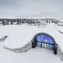Sneklædt bygning med buet indgang oplyst i blåt. Omgivet af et snedækket landskab og fyrretræer, med fjerne huse under overskyet himmel.