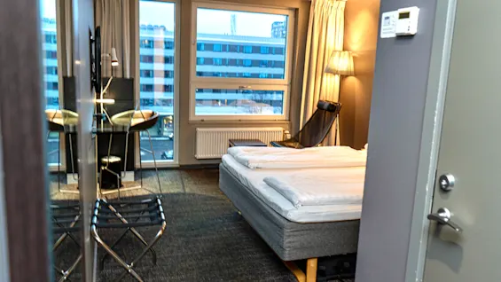Ett modernt hotellrum med en säng, en liten sittgrupp vid fönstret och utsikt över stadens byggnader.