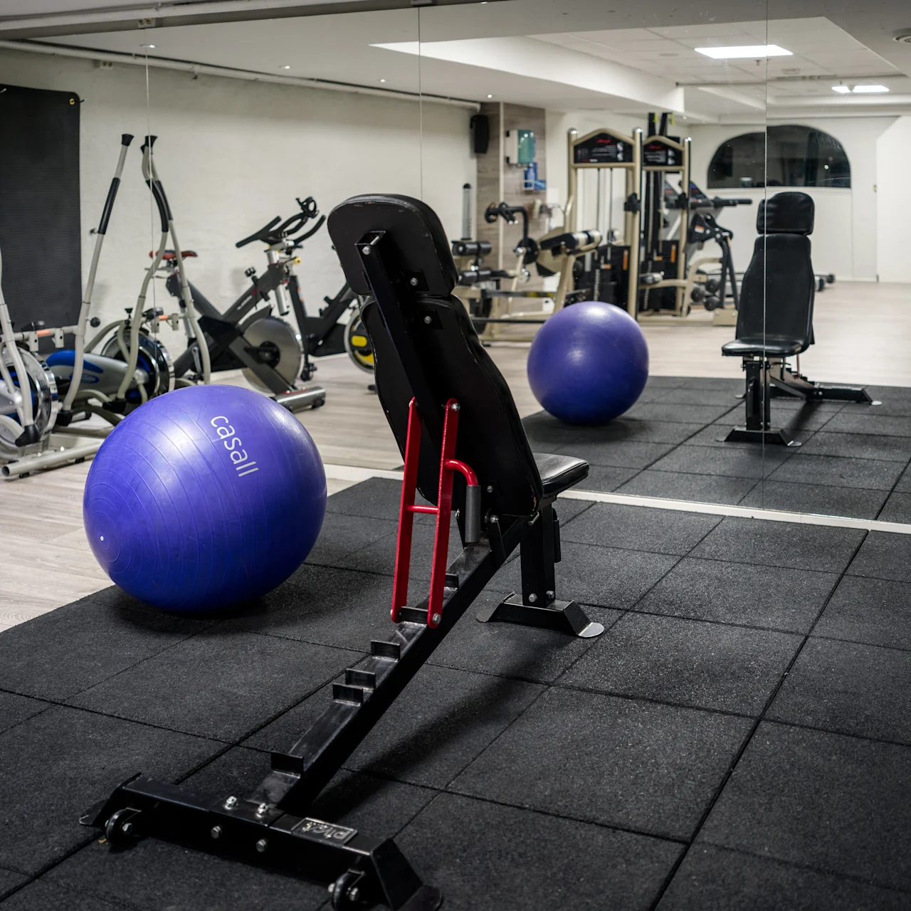 Bench rests on black mat in a gym, with exercise balls and various fitness equipment nearby. The word "Casall" is visible on one blue exercise ball. Mirrors cover one wall.