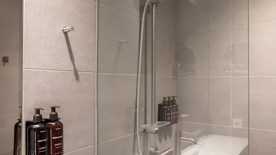 Et moderne bad med grå fliser, en dusj og en vask. To pumpeflasker merket BODY WASH og BODY LOTION er montert på veggen.