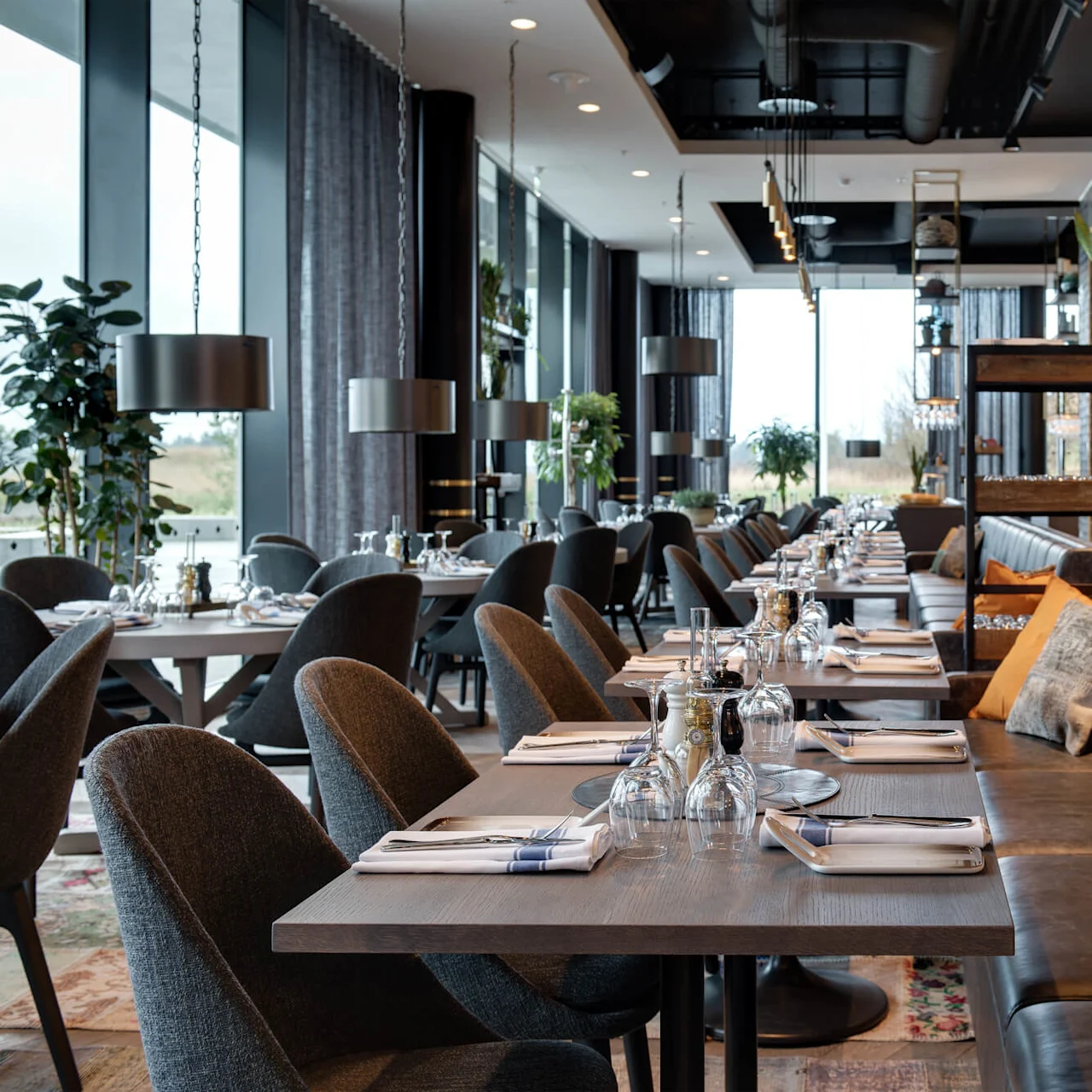 En moderne restaurant har pent arrangerte spisebord med elegante oppsett i et romslig rom. Store vinduer gir naturlig lys, mens dekorative planter og stilige møbler skaper en koselig atmosfære.