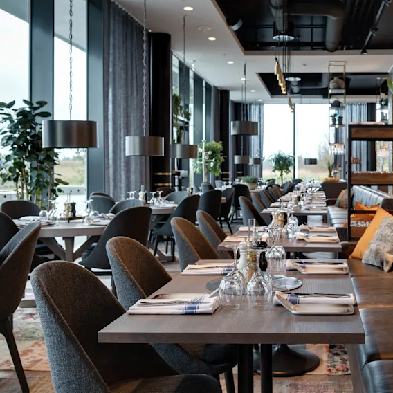 En moderne restaurant har pent arrangerte spisebord med elegante oppsett i et romslig rom. Store vinduer gir naturlig lys, mens dekorative planter og stilige møbler skaper en koselig atmosfære.