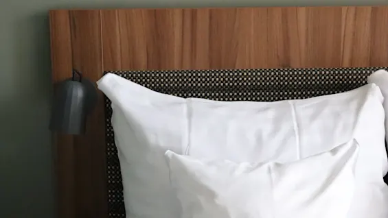 White pillows rest against a wooden headboard in a neatly made bed, beside a black lamp. Above, a colorful abstract artwork decorates the gray wall.