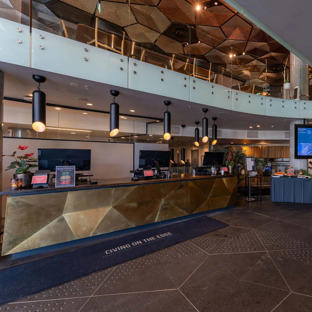 Reception desk with modern lighting fixtures sits beneath a curved glass railing. Monitors display information. Nearby, a table with refreshments is arranged. Floor mat reads “LIVING ON THE EDGE.”