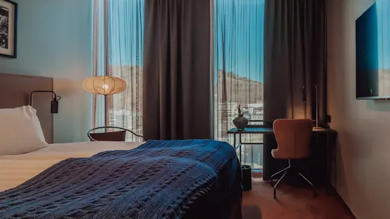 En säng med en blå filt står bredvid en tänd lampa; ett skrivbord med en stol och en vas är nära ett fönster med skira gardiner, med utsikt över stadssiluetten.