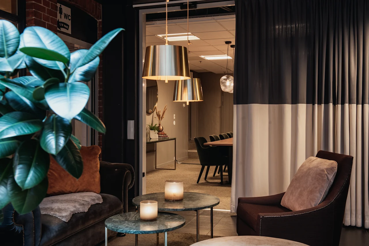 Et stilfuldt loungeområde med komfortable siddepladser og glødende stearinlys, der fører til et moderne mødelokale. WC-skilt synligt.