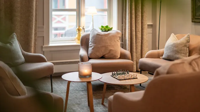 A cozy living room setup features four armchairs around a circular wooden coffee table with a chessboard and candle, framed by a window with drapes and a floor lamp.