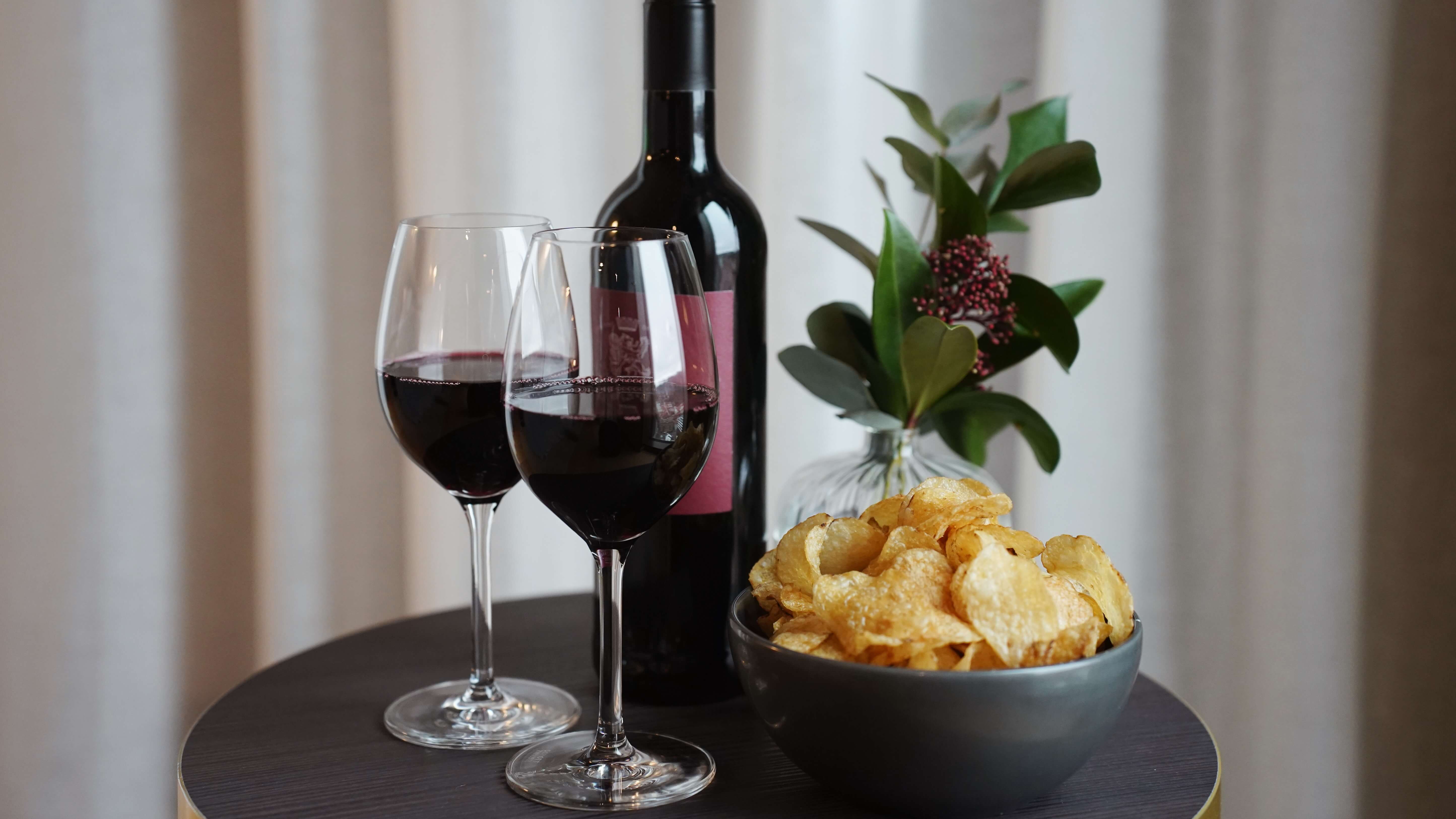 To glass og en flaske rødvin med en bolle potetgull på et bord, med en plante i bakgrunnen.