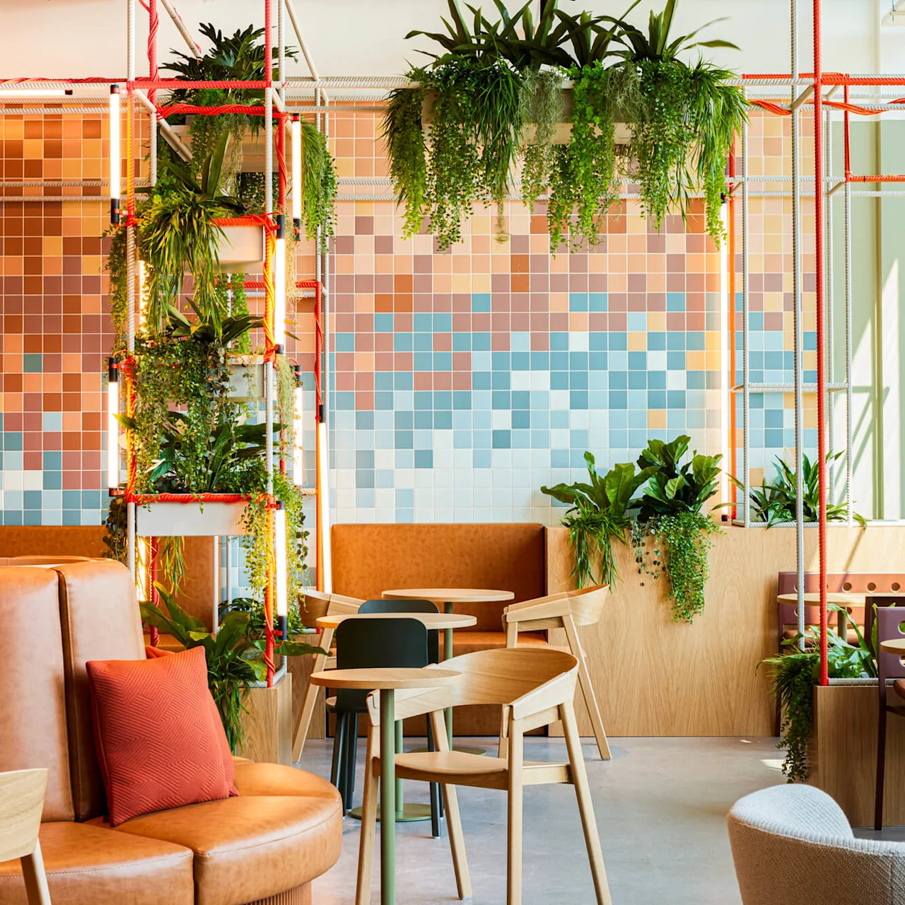 Siddeområde med lædersofaer og træstole omgivet af frodige grønne planter, under et loftsgitter med orange reb. Mosaikvæg danner en farverig baggrund.