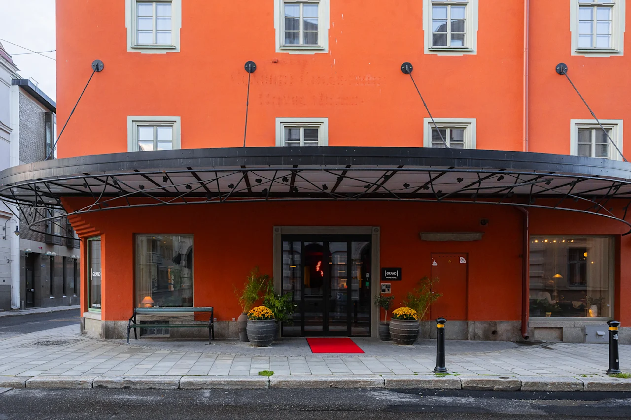 En levende orange bygning med store vinduer har en overdækket indgang med en rød løber. Potteplanter flankerer døren, og skilte siger "hotel." Det ligger på en brostensbelagt gade.