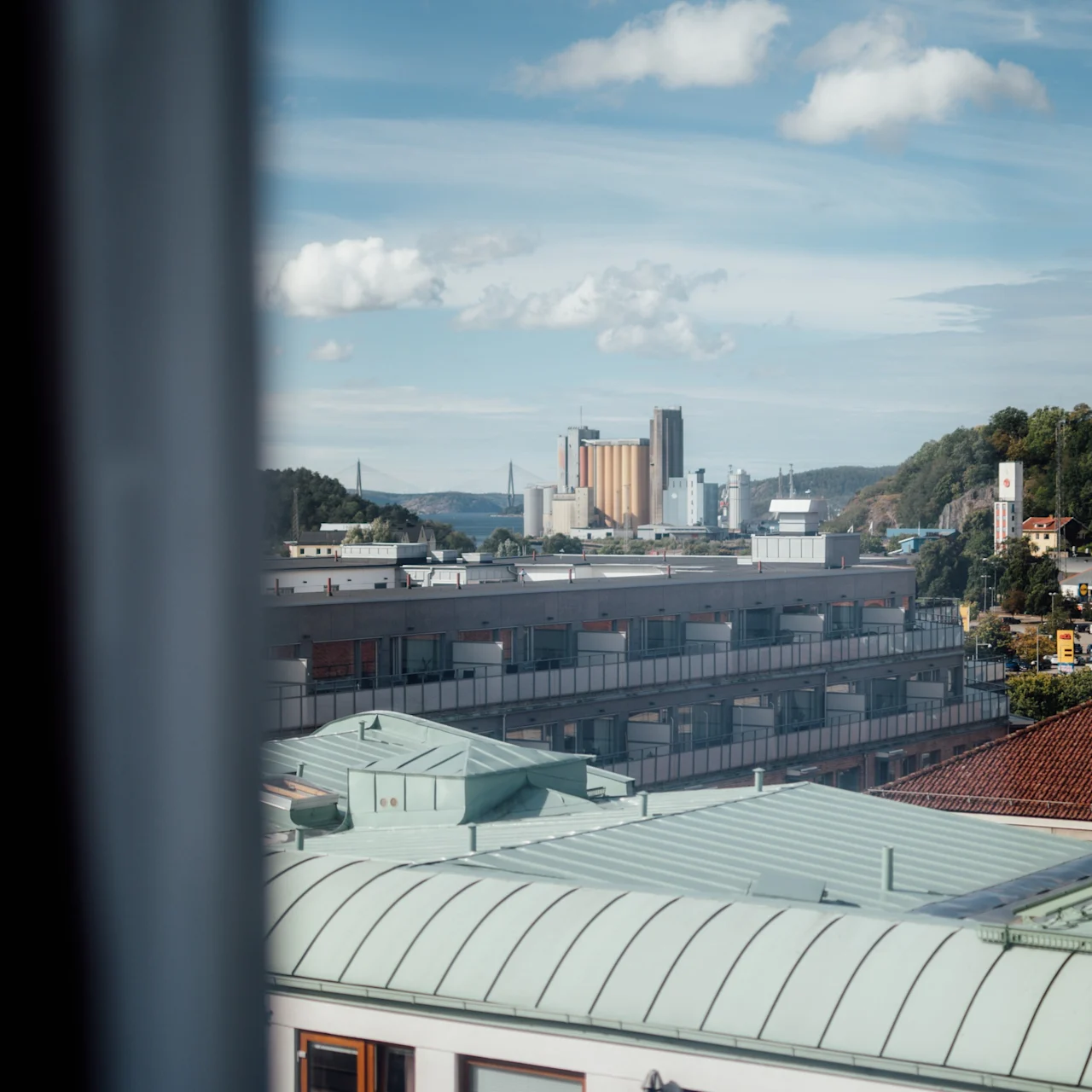 Byggnader med gröna tak ses genom ett fönster, med en avlägsen industriell silhuett och kullar under en delvis molnig blå himmel.
