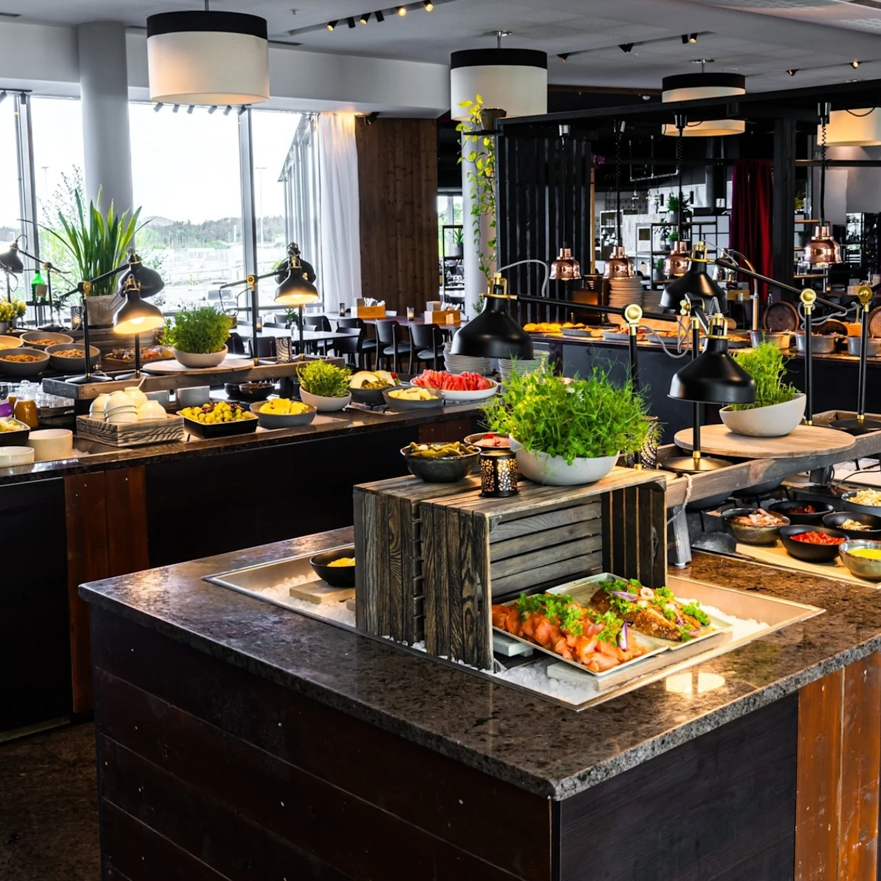 A large buffet table features various dishes including salads, bread, and garnishes. It's surrounded by modern decor with large windows in a well-lit restaurant setting.