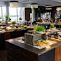 Et stort buffetbord med ulike retter inkludert salater, brød og garnityr. Det er omgitt av moderne innredning med store vinduer i en godt opplyst restaurantsetting.