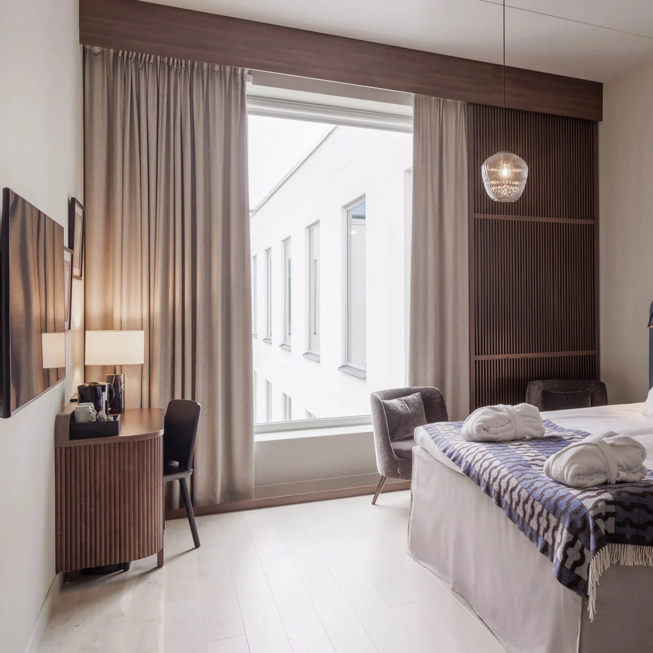 Neatly arranged hotel room with a made bed, side table, and chair, illuminated by natural daylight from a large window.