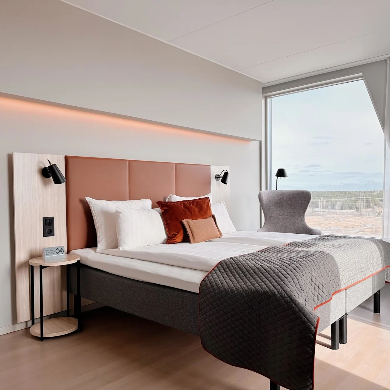 A modern hotel room features a double bed with layered pillows and a quilt, flanked by wall lamps. Large windows offer a scenic view of a forested landscape. 