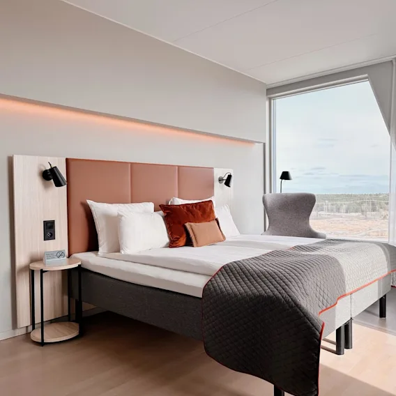 Ett modernt hotellrum har en dubbelsäng med kuddar och filt, omgiven av vägglampor. Stora fönster erbjuder utsikt över ett skogslandskap.