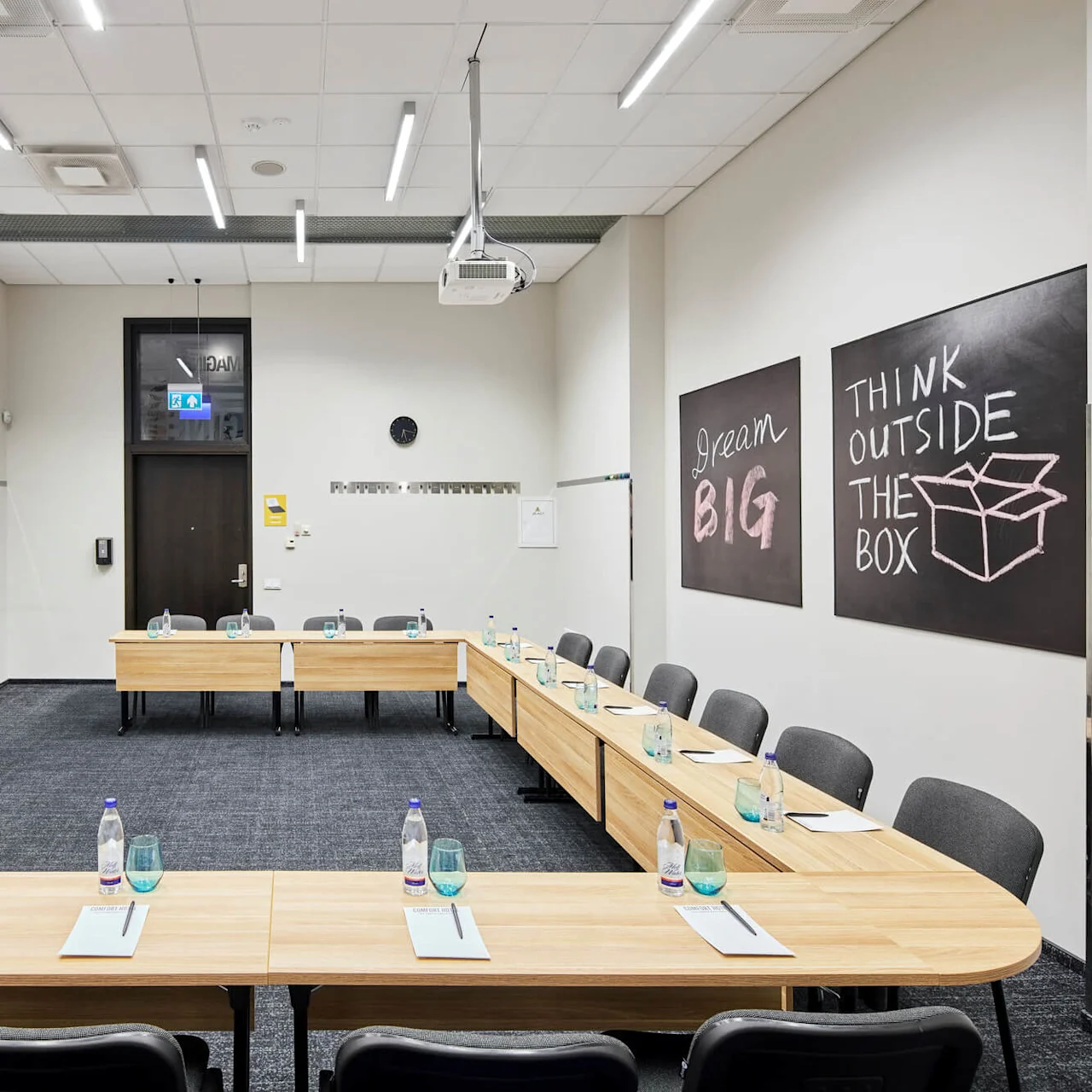 Mötesrum med U-formade trätabeller fyllda med anteckningsblock och vattenflaskor. Två griffeltavlor visar "Dream BIG" och "THINK OUTSIDE THE BOX." Projektor och duk installerade ovanför.