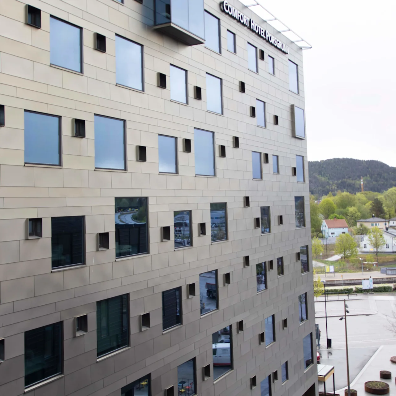 En moderne bygning med mange kvadratiske vinduer, mærket "COMFORT HOTEL PORSGRUNN." Den ligger ved et lille byområde med bjerge i baggrunden.