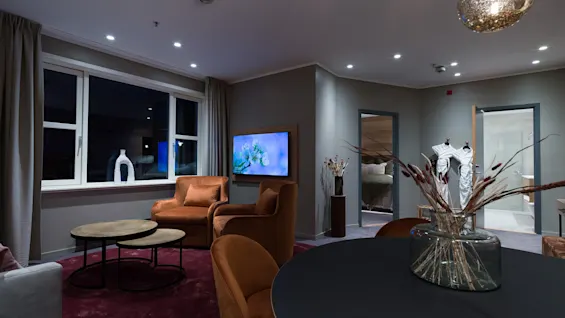 A living room features brown armchairs on a red rug, a circular glass table with dried plants, a wall-mounted TV, and decorative objects near a large curtained window.