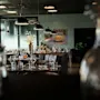 Et moderne restaurantinteriør med dekkede bord, pendellamper og et maleri av en gul bil. En dør med teksten 'HINDEN' er synlig.