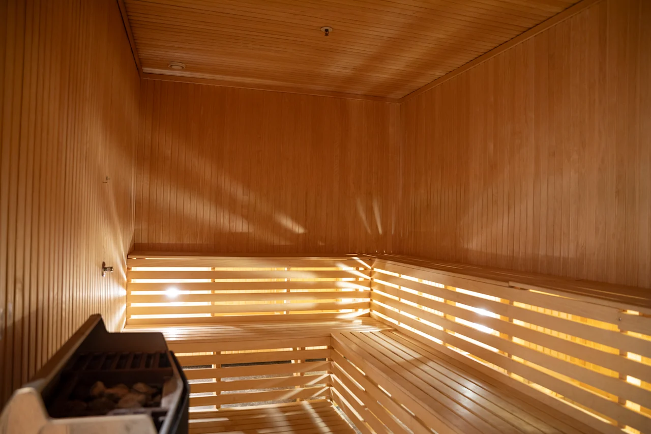 Trebænker beklæder væggene i en varm, lystfyldt sauna. En varmer med sten står i hjørnet og skaber en afslappende atmosfære med sine træpaneler.