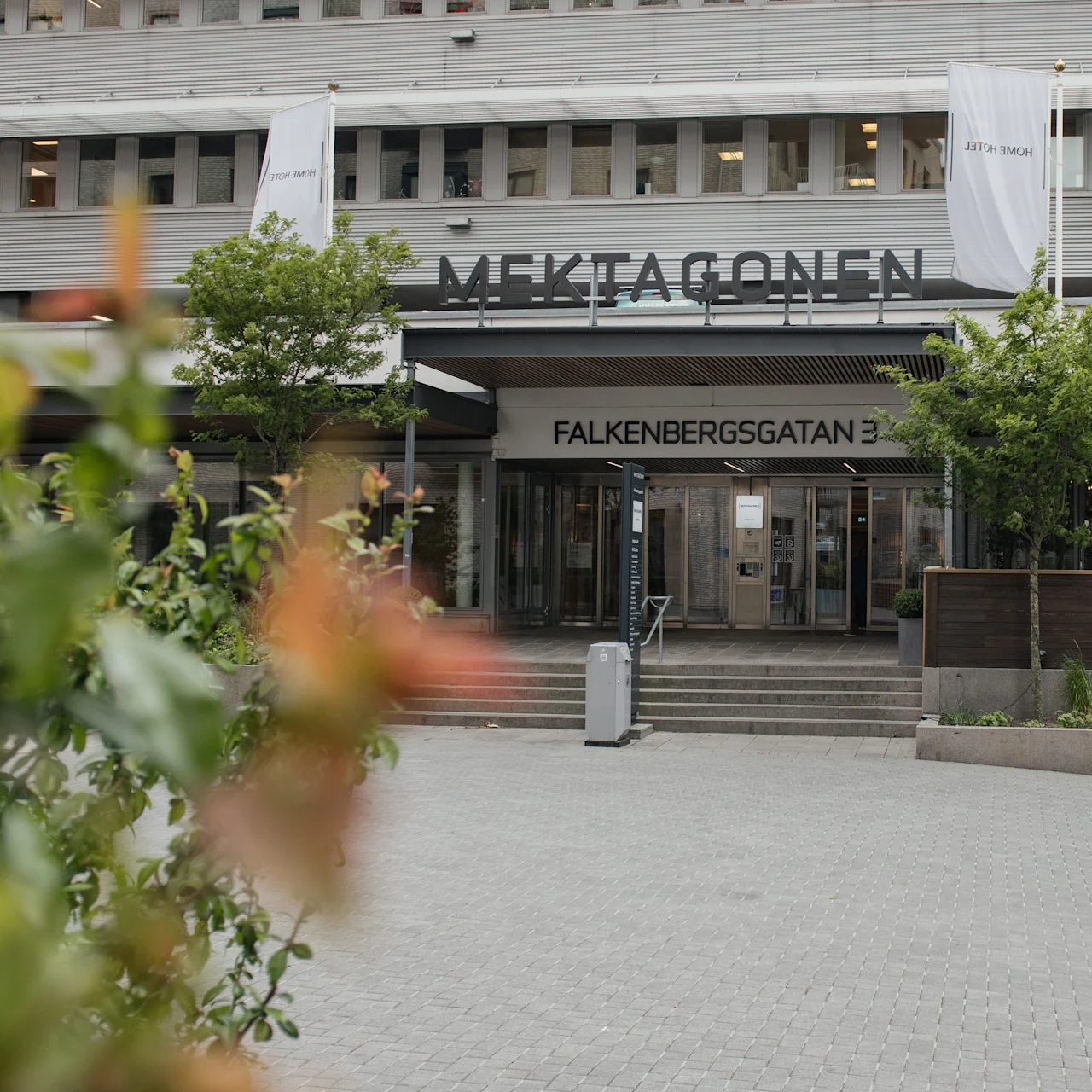 Building entrance displaying "MEKTAGONEN" and "FALKENBERGSGATAN 3" above. Surrounded by trees, bushes, and paved courtyard. White banners hang on the facade.