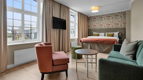 Ett modernt hotellrum med en säng och blommigt huvudgavel, fåtölj, soffa, och en väggmonterad TV, samt stadsutsikt från fönstret.