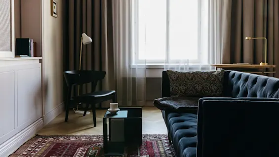 En plysjsofa i mørk farge står i forgrunnen med en mønstret pute, ved siden av et vindu med lette gardiner. Et salongbord har en kopp. En stol og lampe står i nærheten. Bøker og et bilde pryder vegghyllene.