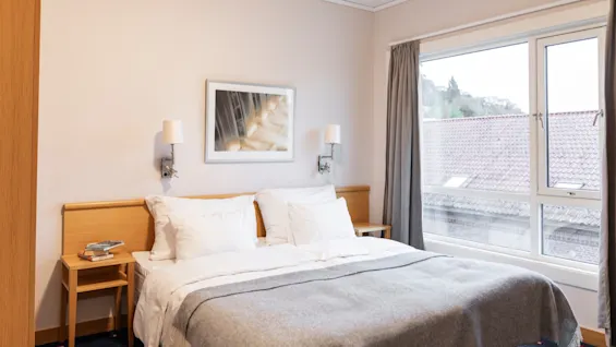 Et koselig hotellrom med en komfortabel seng, to nattbord og et stort vindu med utsikt over hustak.