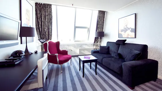 Modern living room with a black couch, red chair, and large windows. Art portraits and a flat-screen TV adorn the walls.