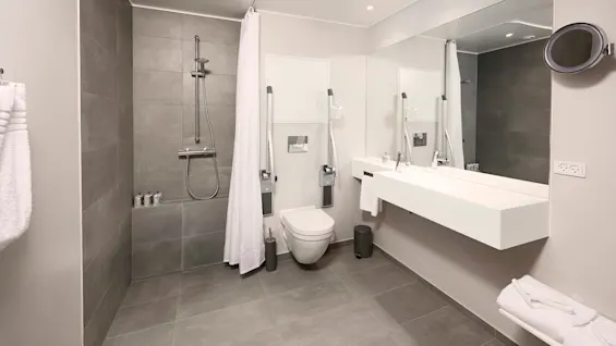 Nykyaikaisessa kylpyhuoneessa on kävelysuihku, seinään asennettu wc tukikaiteilla, suuri peili kaksoispesualtaan yläpuolella, pyyhetelineet ja minimalistinen muotoilu neutraaleissa sävyissä.