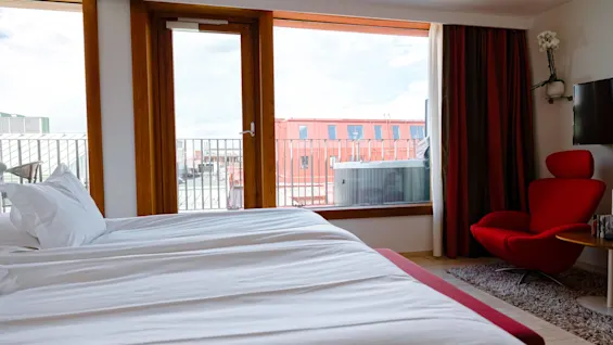 A neatly made bed faces a large window with a city view. A red armchair sits beside a television in a modern, clean room setting.