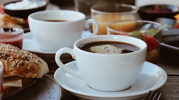 A close-up of a coffee cup, steaming on a saucer, amidst a breakfast setting with various foods and beverages on a wooden table.