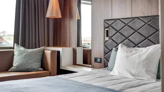 A modern bedroom with a neatly made bed, diamond-patterned headboard, wood paneling, a pendant lamp, and a long curtain. Contemporary, cozy atmosphere.