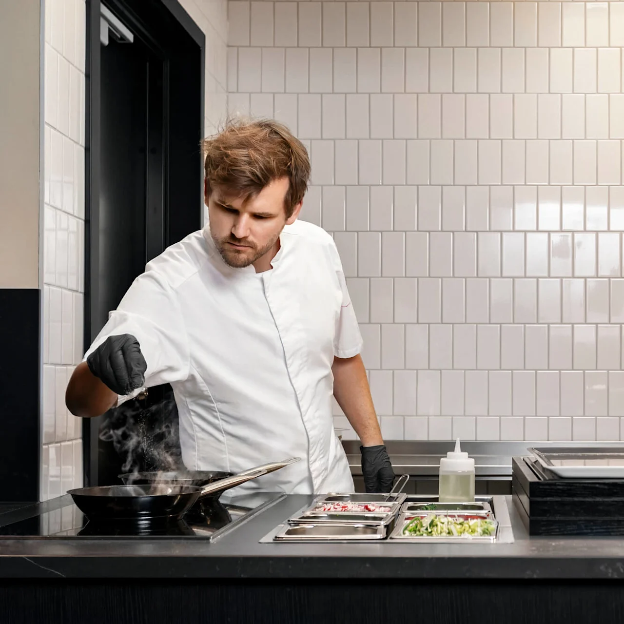 Kocken strör kryddor från vänster hand i en ångande panna på spisen, omgiven av ingredienser och köksredskap, kaklad vägg med ljus lampa i modernt professionellt kök.