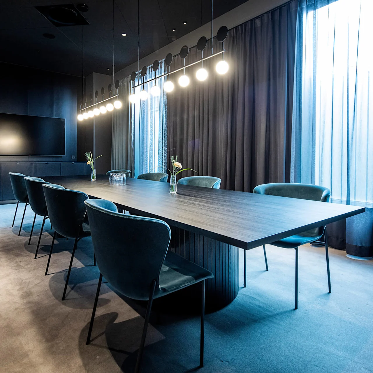 Ett långt konferensbord med blå stolar är placerat i ett rum med en modern ljusarmatur ovanför. Gardiner kantar väggen, och en plattskärms-TV är monterad mittemot.