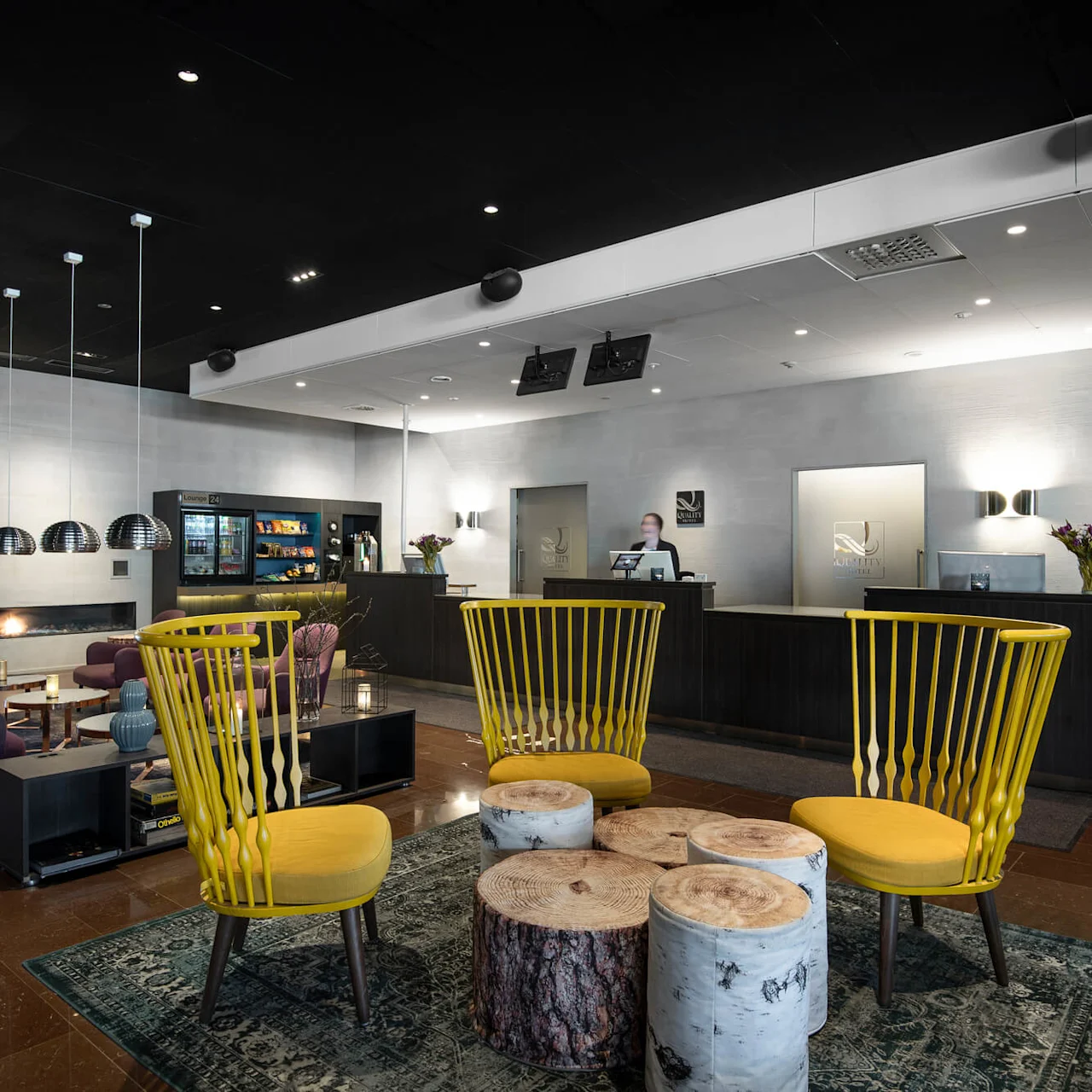 Receptiondisk med en person som arbetar, omgiven av stilfulla möbler i en modern lobby. Gula stolar och stockbord kompletterar mörka soffor, växter och hängande lampor. Text: "QUALITY HOTEL."
