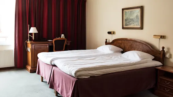 En dobbeltseng med hvitt sengetøy og en trehodegavl står på et hotellrom. Mørkerøde gardiner dekker vinduet, og et skrivebord med en lampe er i nærheten.
