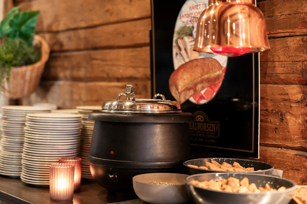 Et rustikt buffetbord med en suppevarmer, stablede tallerkener og madskåle, oplyst af varm belysning. KARET TIL SANDESJORD.