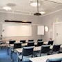 Et lyst, tomt konferencelokale med rækker af borde og stole vendt mod en whiteboardtavle og projektor, klar til et møde eller en præsentation.