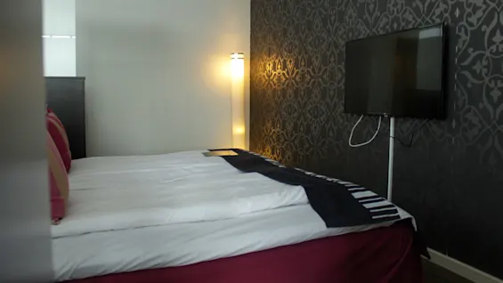 En pent oppredd seng med dekorative puter, flankert av en stående lampe og en veggmontert TV, i et rom med mønstret tapet.