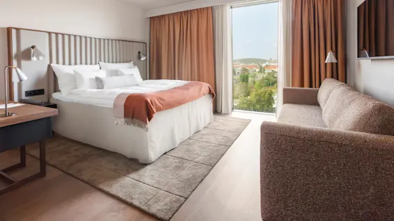 En säng med vita lakan och en orange pläd står vid ett fönster med utsikt över staden. I närheten finns en brun soffa och en skrivbordslampa i ett modernt, ljust rum.