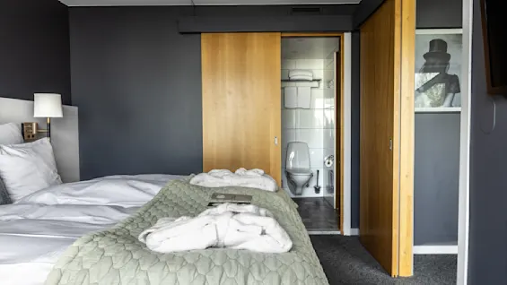 Ett mysigt hotellrum med en snygg bäddad säng, mjukt grön filt och morgonrock leder till ett badrum genom skjutdörrar. En lampa ger värme och ett modernt konstverk smyckar väggen.
