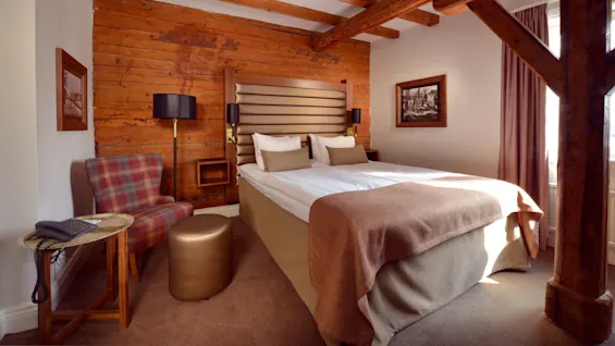 A cozy bedroom with a double bed, wooden walls, and a checkered chair, illuminated by natural light from an adjacent window.
