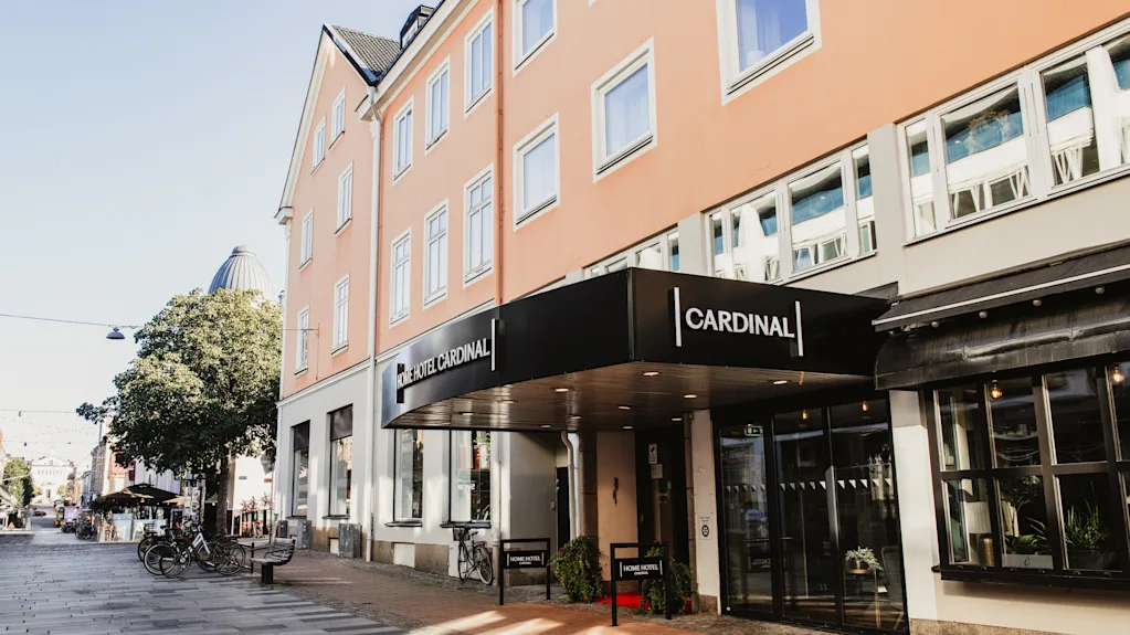 Hotellin sisäänkäynti aurinkoisella kadulla, jossa on musta markiisi, jossa lukee HOTEL CARDINAL. Polkupyöriä on pysäköity lähelle.