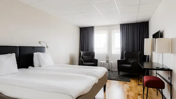 Två prydligt bäddade enkelsängar står i ett minimalistiskt hotellrum, med två fåtöljer bredvid ett runt bord, gardiner över ett fönster och en TV på väggen.