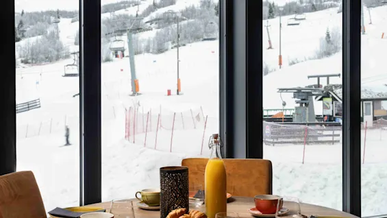 Et rundt træbord dækket med morgenmadsvarer, herunder kopper og croissanter, i en hyggelig spisestue. Store vinduer viser en snedækket skiløjpe udenfor.