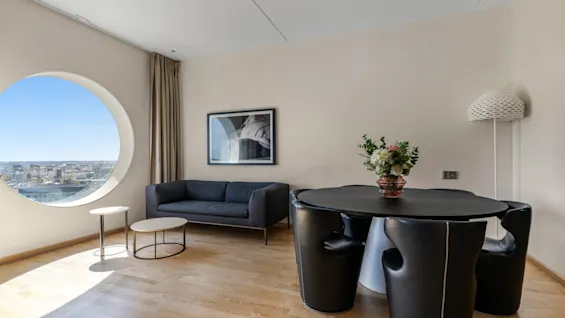 En moderne hotelsuite med rundt vindue, byudsigt, komfortabel sofa og spiseområde med blomsteropsats.