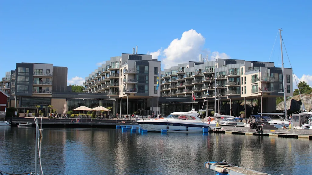 Modern multi-story buildings stand by a marina with several anchored boats. Outdoor seating areas with umbrellas and flags are visible under a clear blue sky.