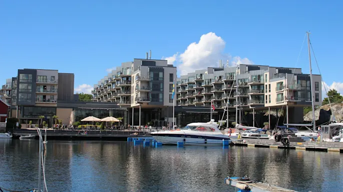 Moderne leilighetsbygninger står ved en marina med flere fortøyde båter. Uteområder med parasoller og flagg ses under en klar blå himmel.